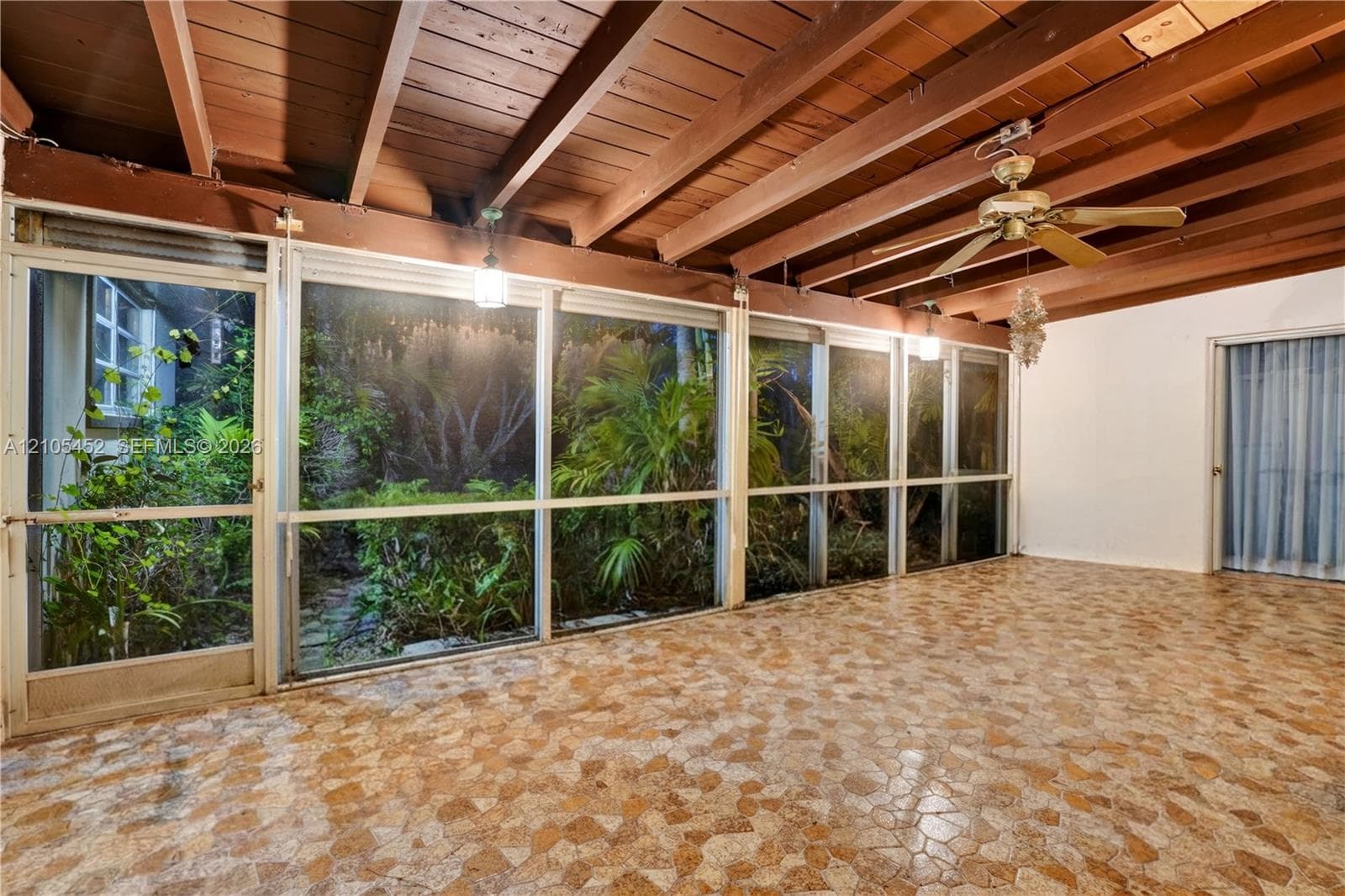 Enclosed patio featuring a natural stone-patterned floor, exposed wood-beam ceiling, floor-to-ceiling screened panels, and a central ceiling fan