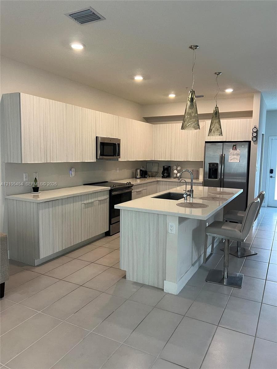 Kitchen featuring light wood-finish cabinetry, stainless steel appliances, a center island with sink, recessed lighting, and tile flooring