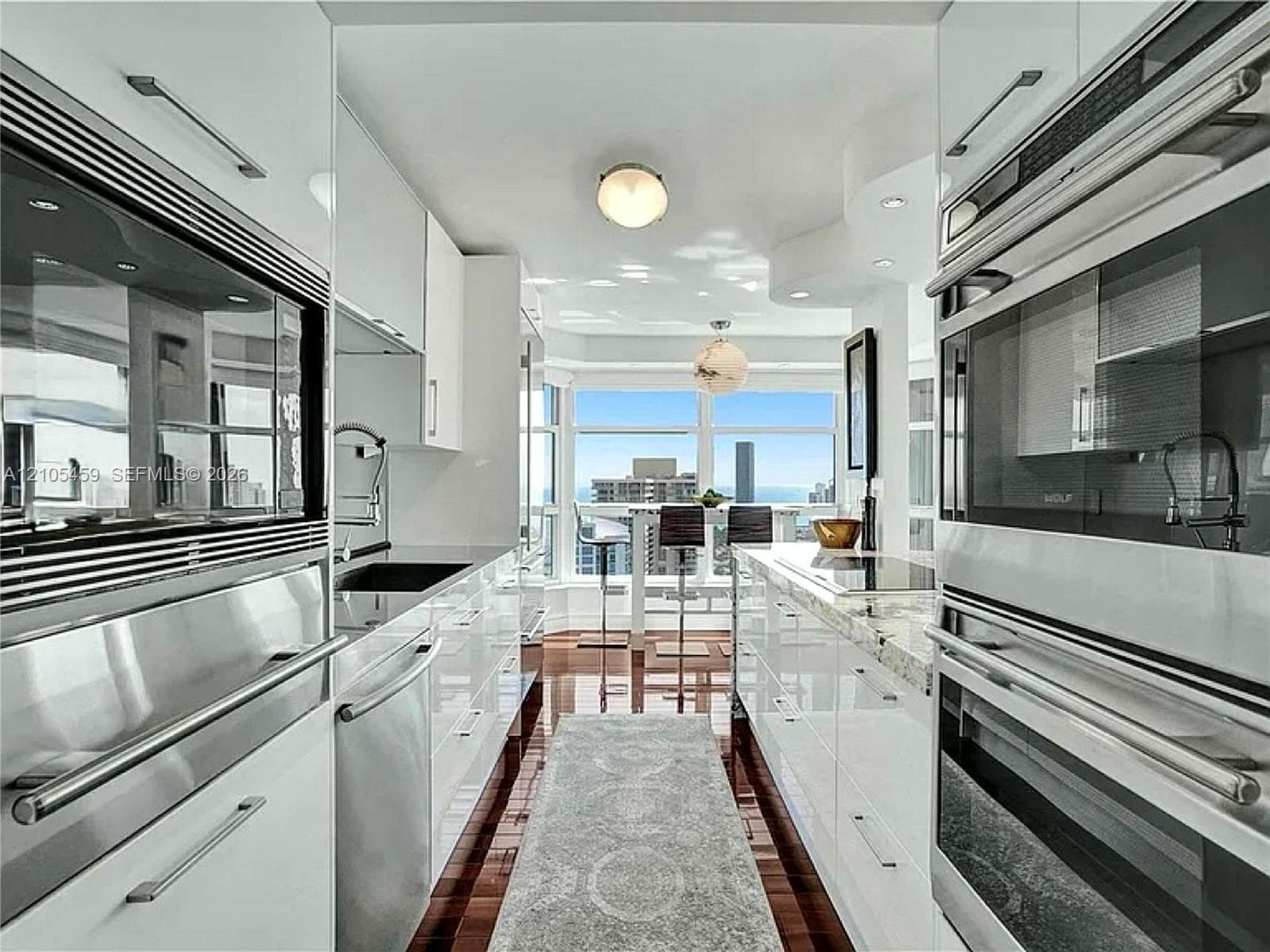 Galley kitchen featuring hardwood flooring, white high-gloss cabinetry, integrated stainless steel appliances, and stone countertops