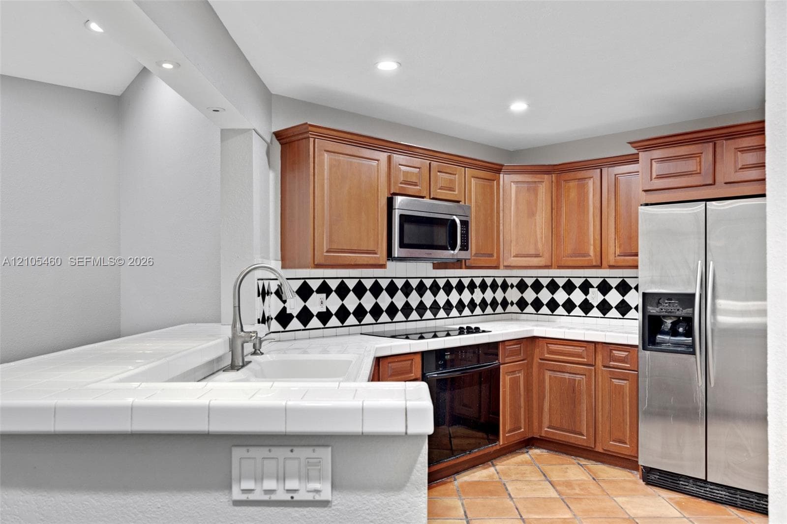 Kitchen featuring wood cabinetry, stainless steel appliances, a black and white diamond-patterned tile backsplash, white tile countertops, and terracotta-style floor tiles