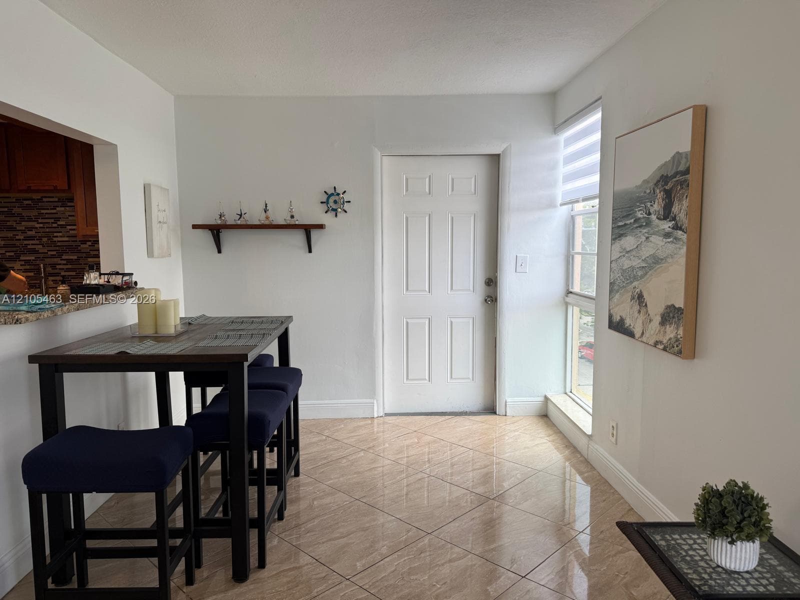 Light beige tile flooring, white paneled door, kitchen pass-through window with granite-style countertop, and a built-in wall shelf