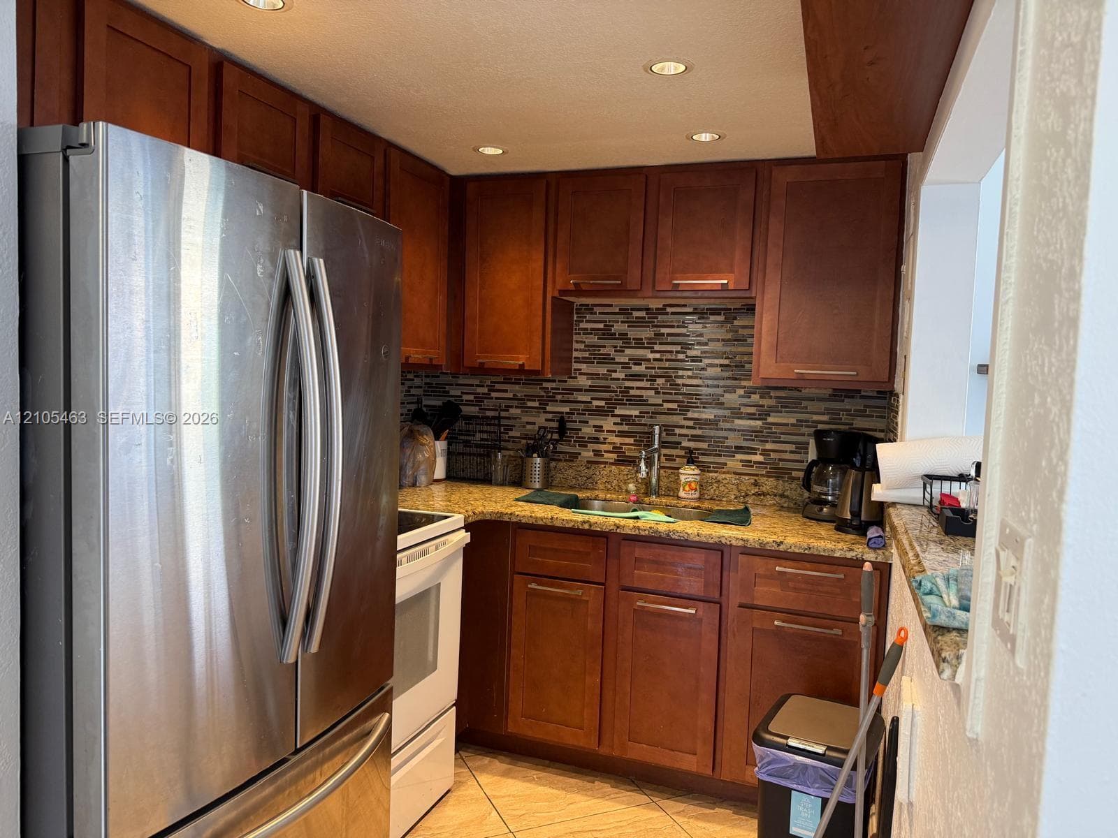 Kitchen featuring wood cabinetry, mosaic tile backsplash, granite-style countertops, stainless steel refrigerator, and recessed lighting
