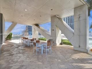 Spacious outdoor patio with tiled flooring, structural concrete columns, a glass railing, and distant water scenery