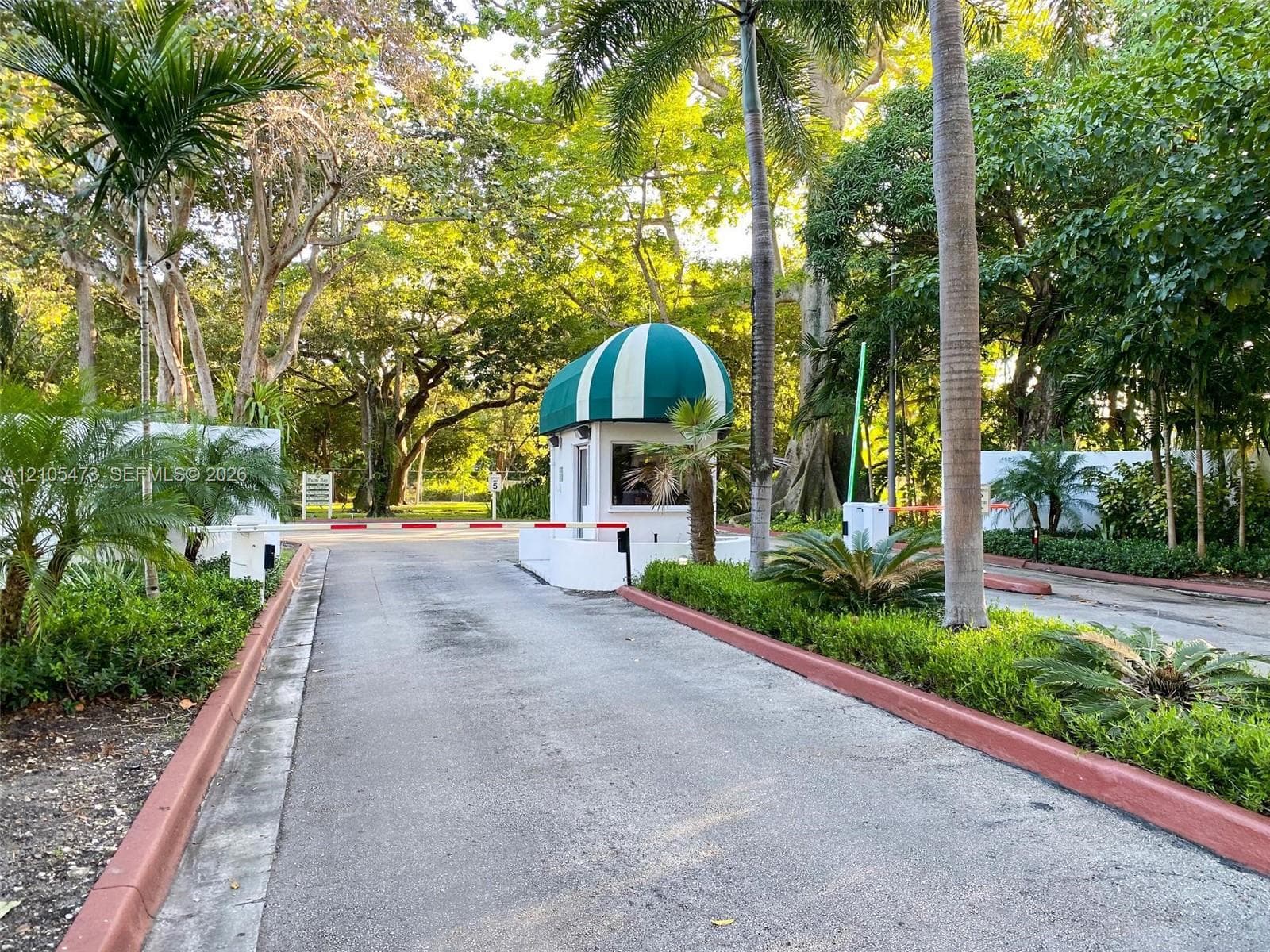 Gated entrance featuring a guardhouse with a green and white striped awning, controlled access barrier, and mature tropical landscaping