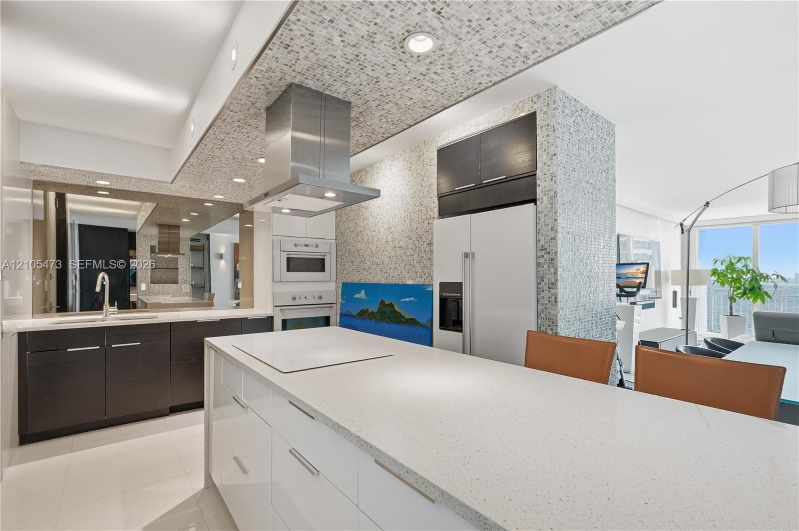 Kitchen featuring a central island with an integrated cooktop, white countertop, dark wood-finish cabinetry, stainless steel range hood, and integrated wall oven
