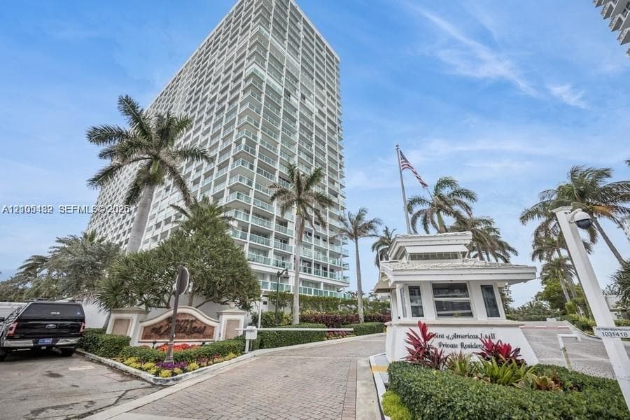 High-rise residential exterior featuring a gated entry with a dedicated security kiosk, paver driveway, and professional landscaping with mature palm trees