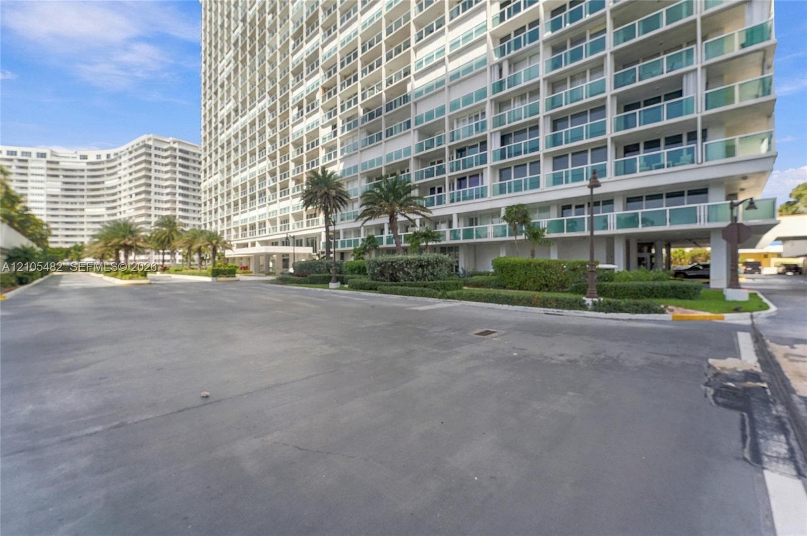 High-rise building exterior featuring glass balcony railings, white structural columns, paved driveway, and mature palm tree landscaping