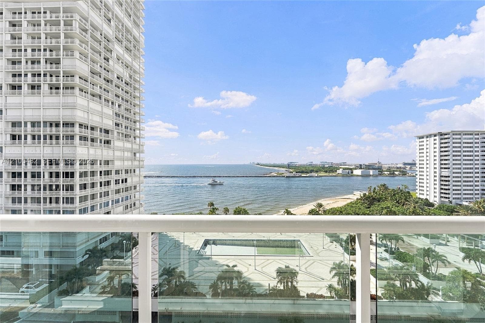 Balcony with a white metal railing and glass paneling, overlooking the ocean and a paved plaza with palm trees