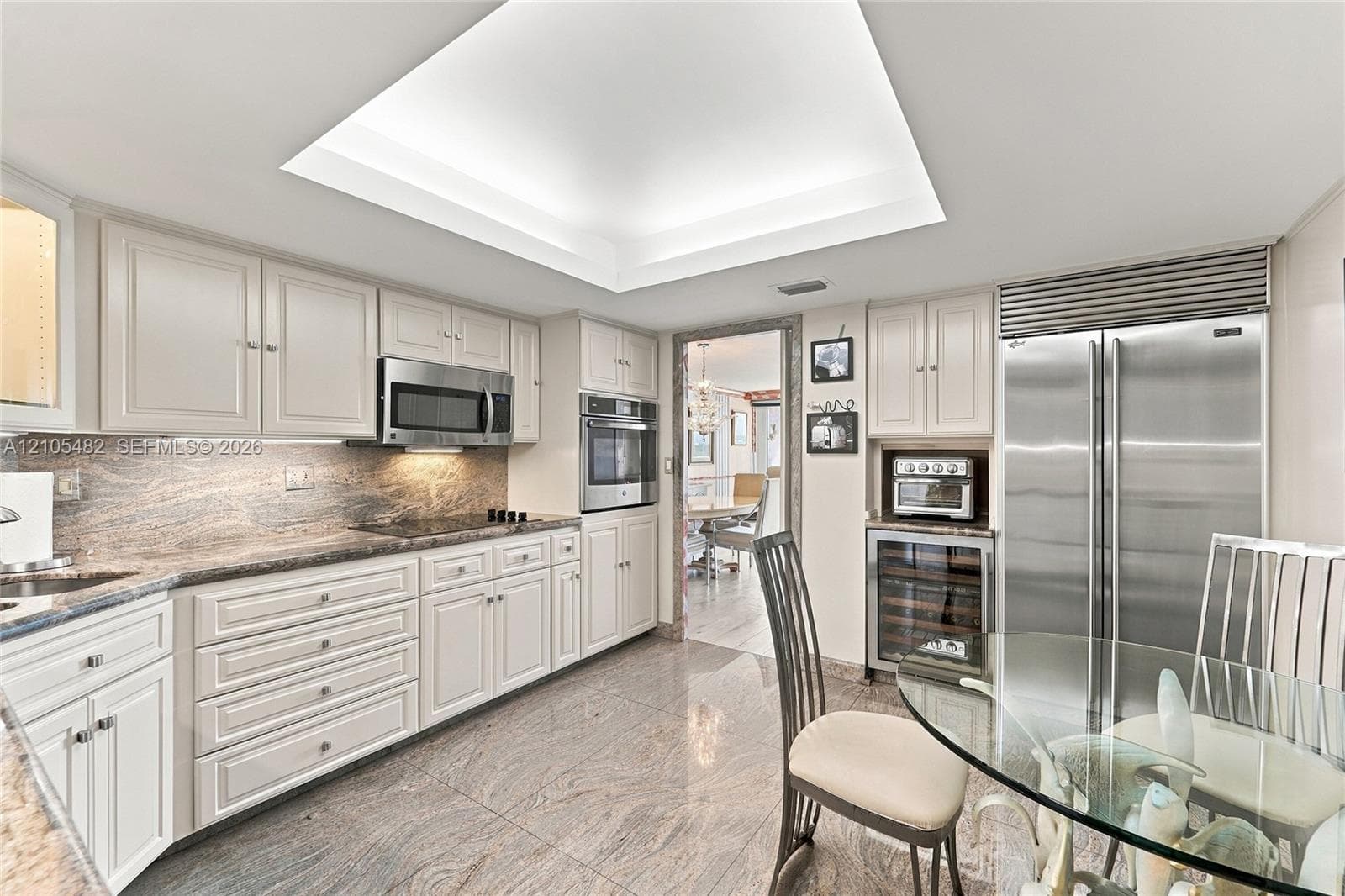 Kitchen featuring light-toned cabinetry, stone countertops with matching backsplash, stainless steel appliances, a built-in wine cooler, and a recessed tray ceiling