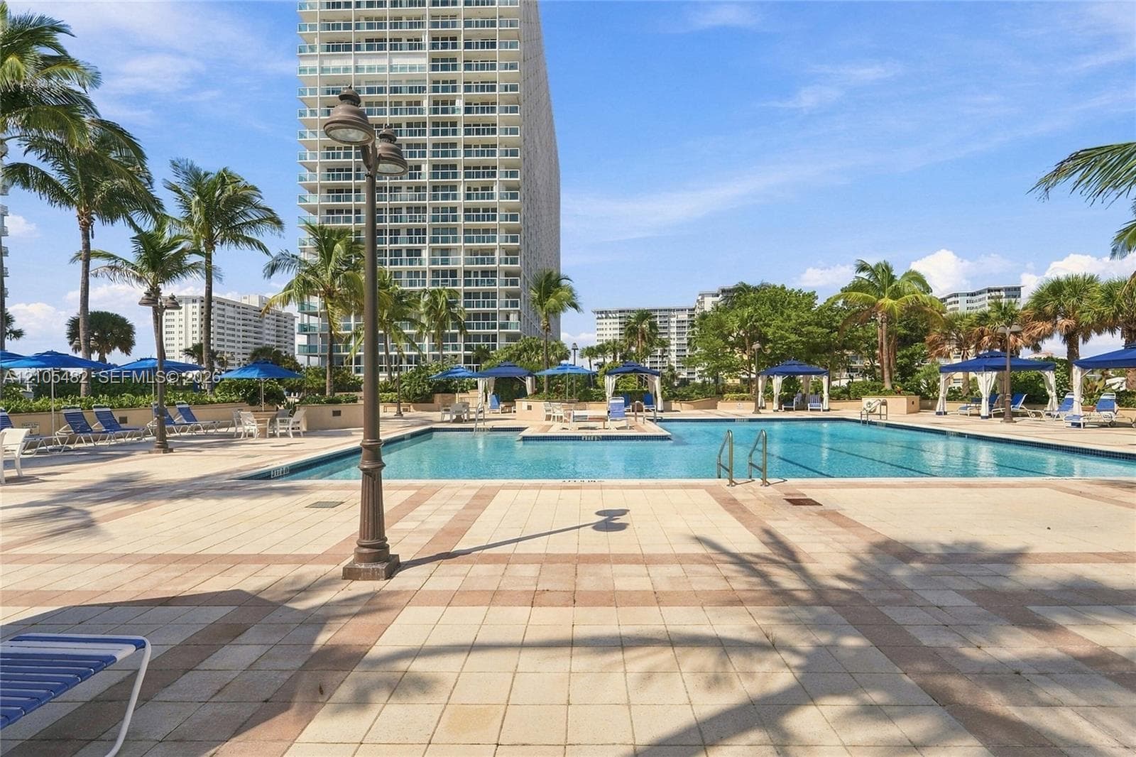 Swimming pool with tiled decking, poolside cabanas, metal post lamps, and landscaping featuring mature palm trees