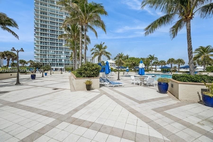Pool deck featuring light-toned stone paving, geometric tile patterns, perimeter landscaping with mature palms, and integrated low-profile retaining walls