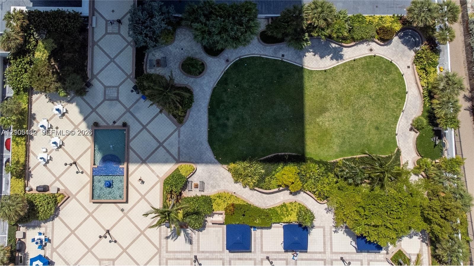Aerial view of outdoor grounds featuring a landscaped lawn, stone-paved walkways, a pool with a tile-accented water feature, and established tropical vegetation