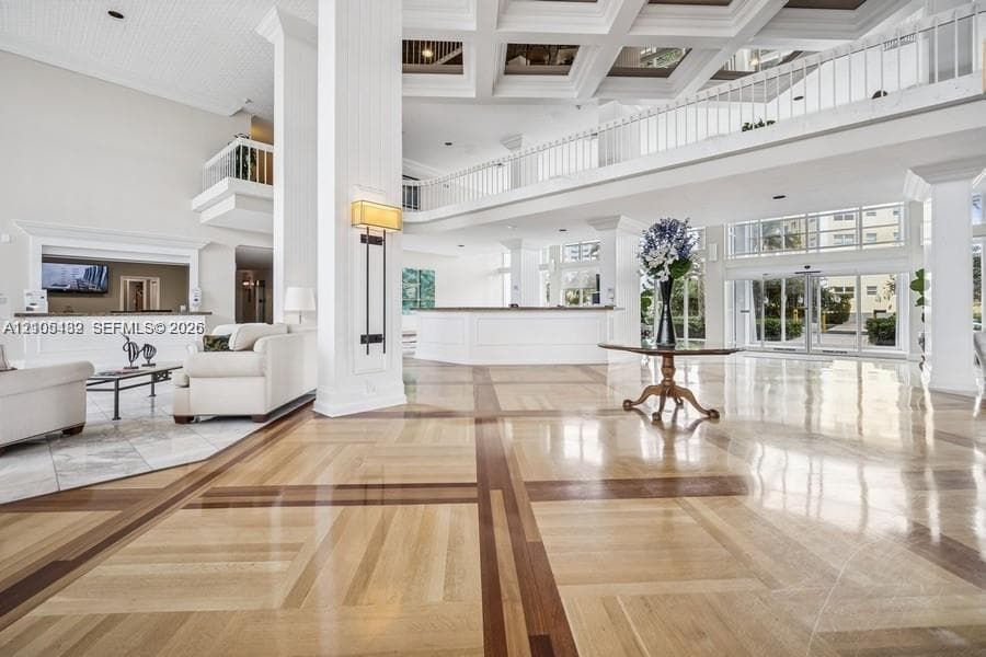 Grand lobby featuring double-height coffered ceilings, multi-tonal inlay wood-finish flooring, a mezzanine level with white balustrade railings, and expansive glass-paneled entry doors