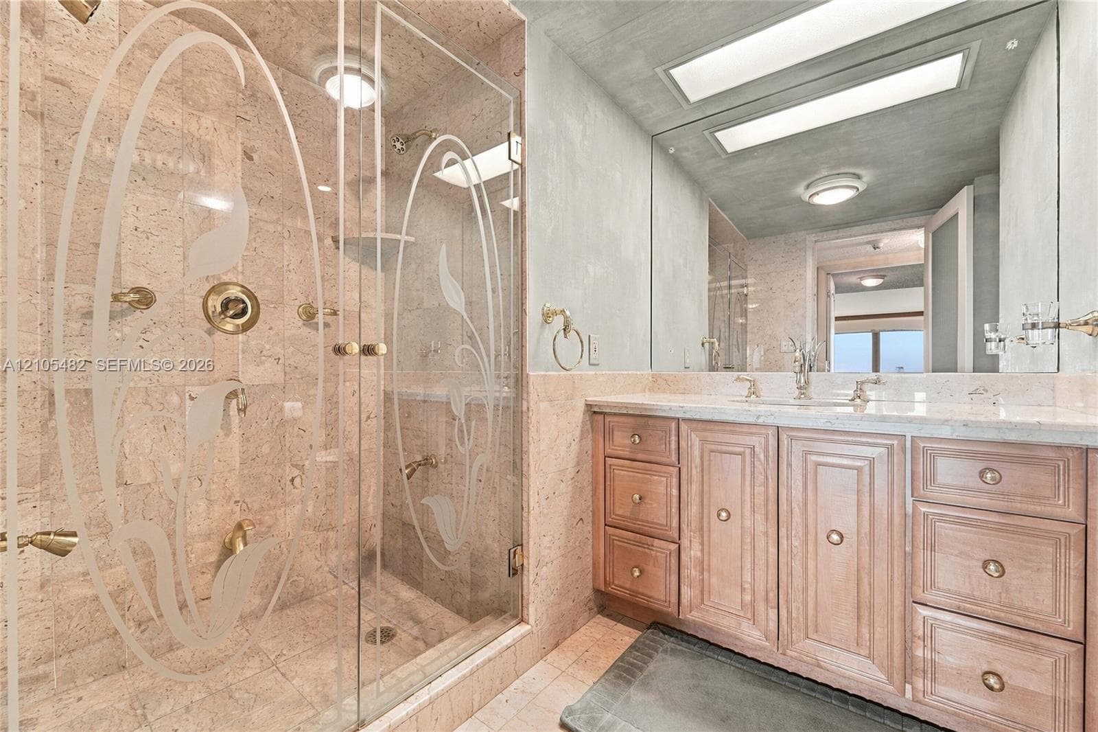 Bathroom featuring a walk-in shower with etched glass doors, stone wall cladding, gold-tone fixtures, wood-finish cabinetry, and an expansive wall-to-wall mirror