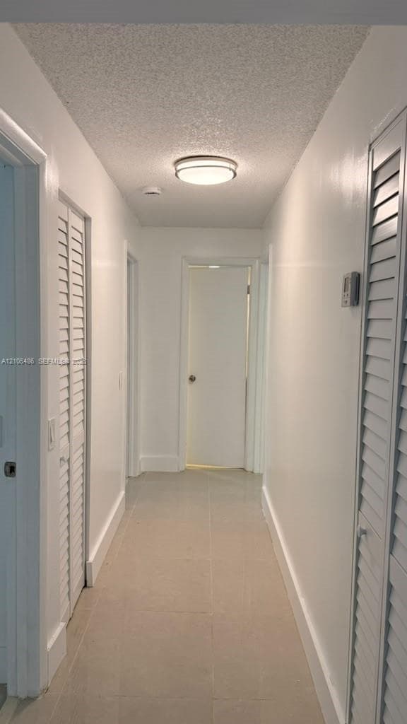 Hallway featuring neutral large-format tile flooring, textured ceiling, dual white louvered closet doors, and a flush-mount circular light fixture