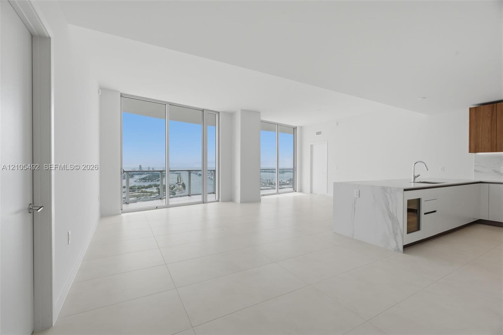 Open-concept living space featuring floor-to-ceiling windows, large-format tile flooring, a kitchen island with stone-finish waterfall countertop, and wood-finish upper cabinetry