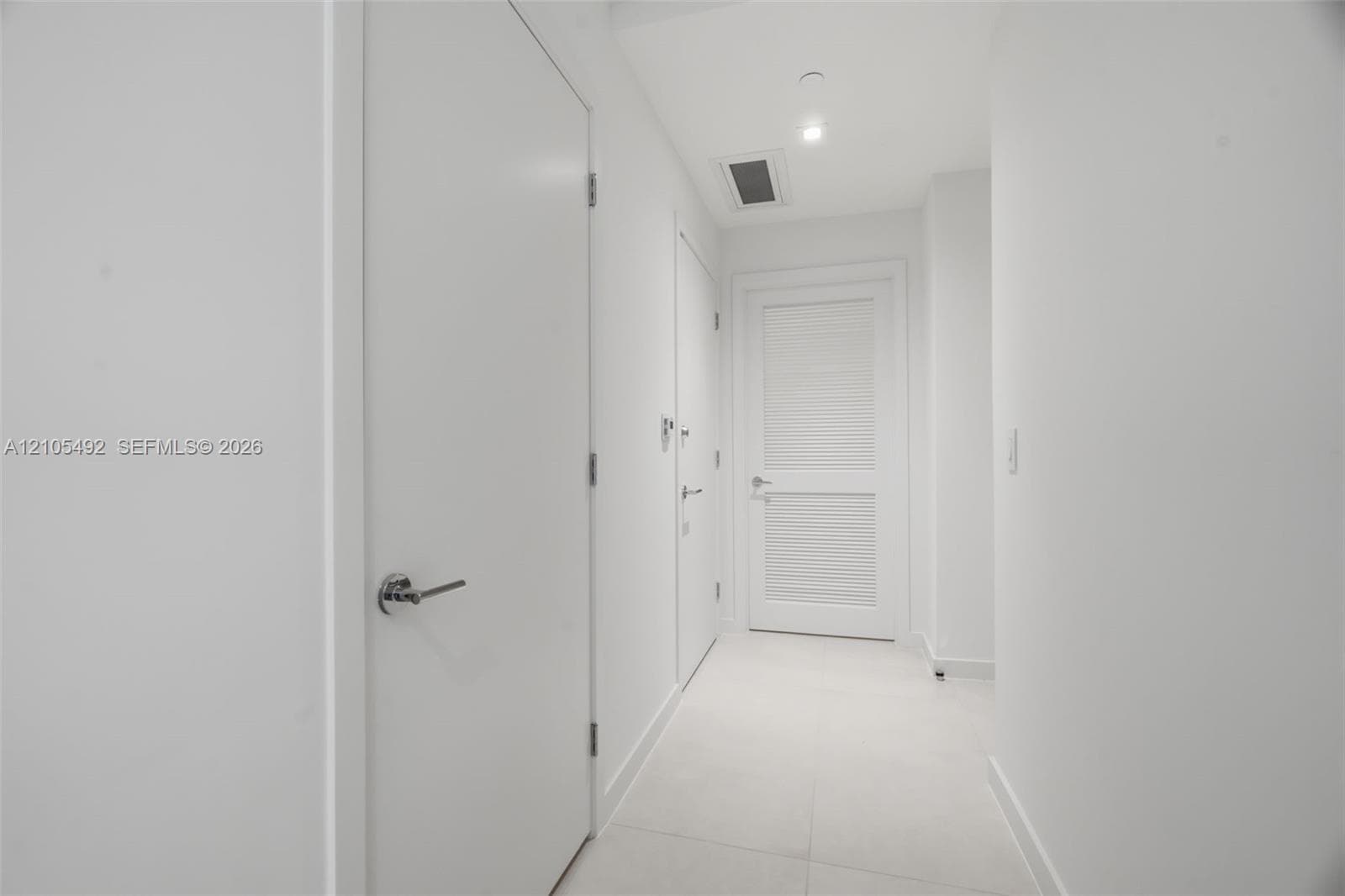 Interior corridor featuring smooth white walls, large-format tile flooring, recessed ceiling lighting, and solid-core doors with brushed-metal lever hardware