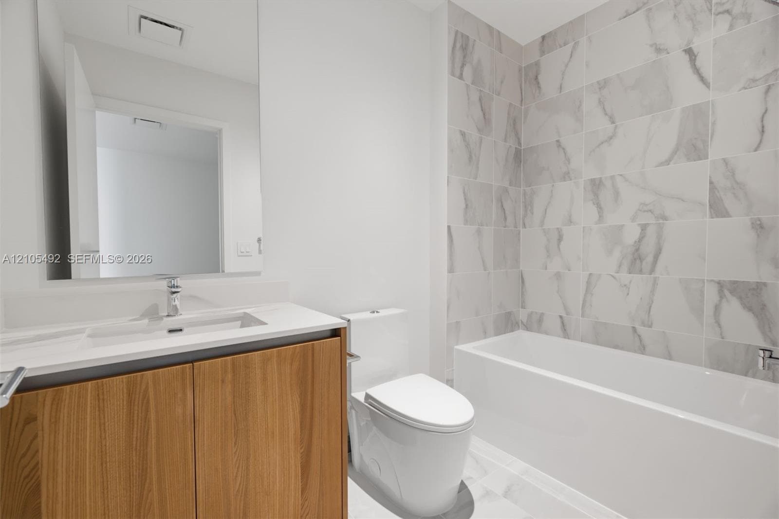 Bathroom featuring a wood-finish vanity with a rectangular sink, stone-surface countertop, floor-to-ceiling tile surround, and a deep soaking tub
