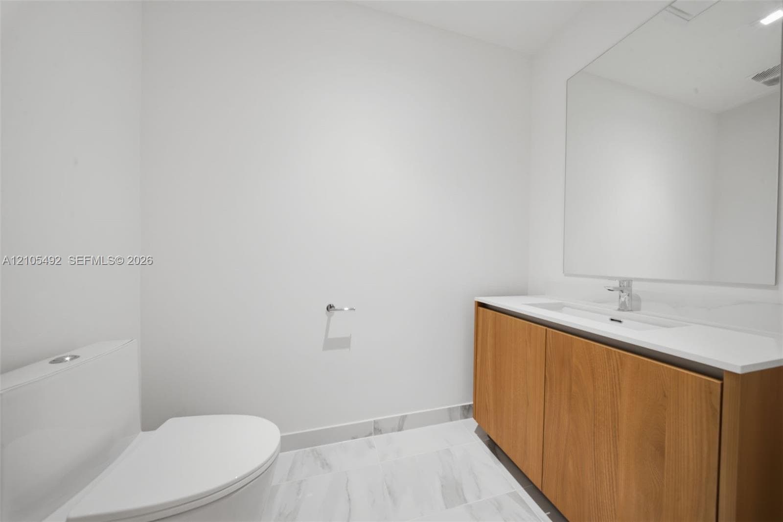 Modern vanity featuring wood-finish cabinetry, a white countertop with an integrated sink, a large frameless mirror, and light-toned tile flooring
