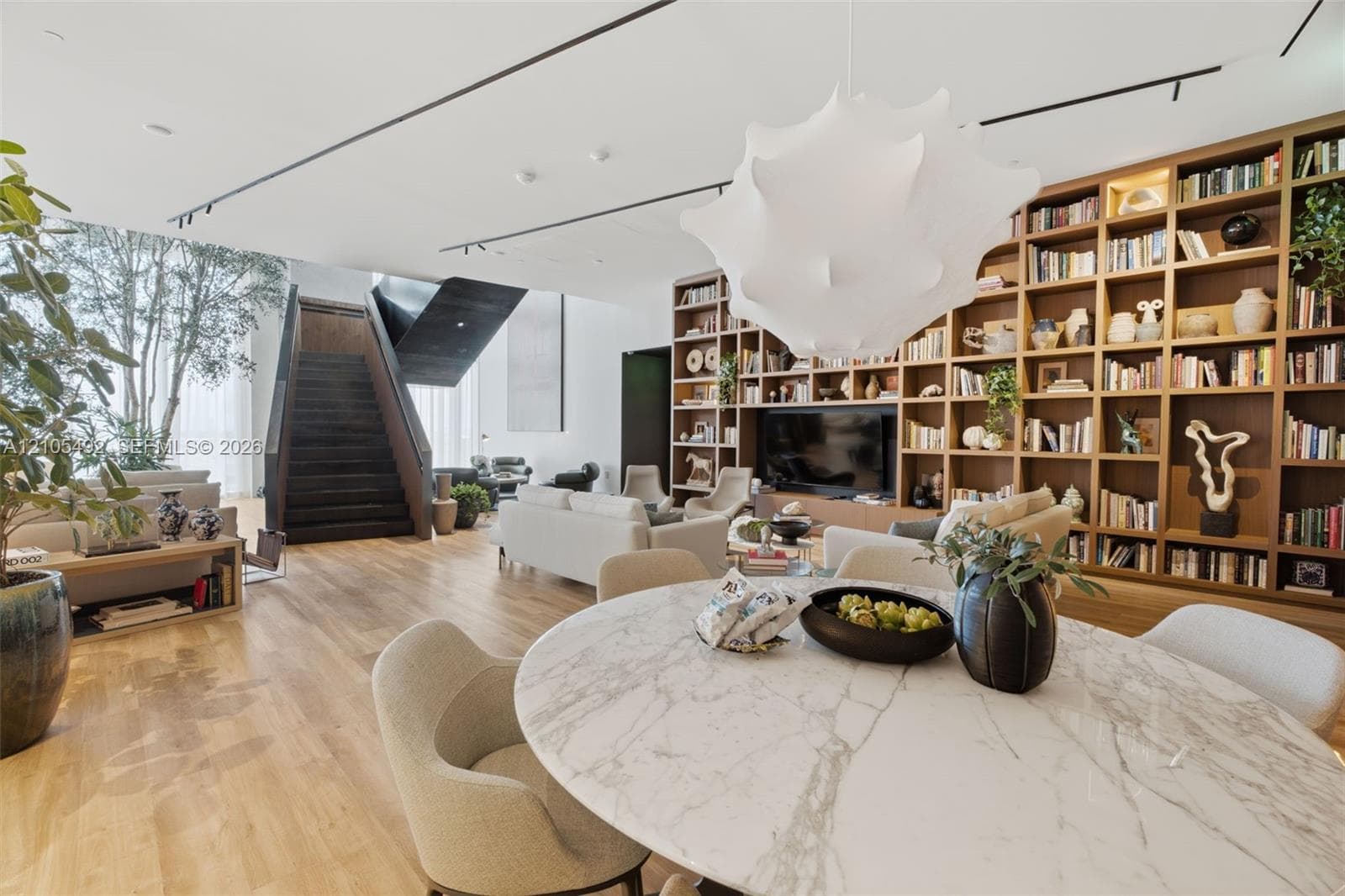 Open-concept interior featuring wood-finish flooring, a floor-to-ceiling built-in wooden bookcase, an architectural staircase with dark metal detailing, and modern track lighting