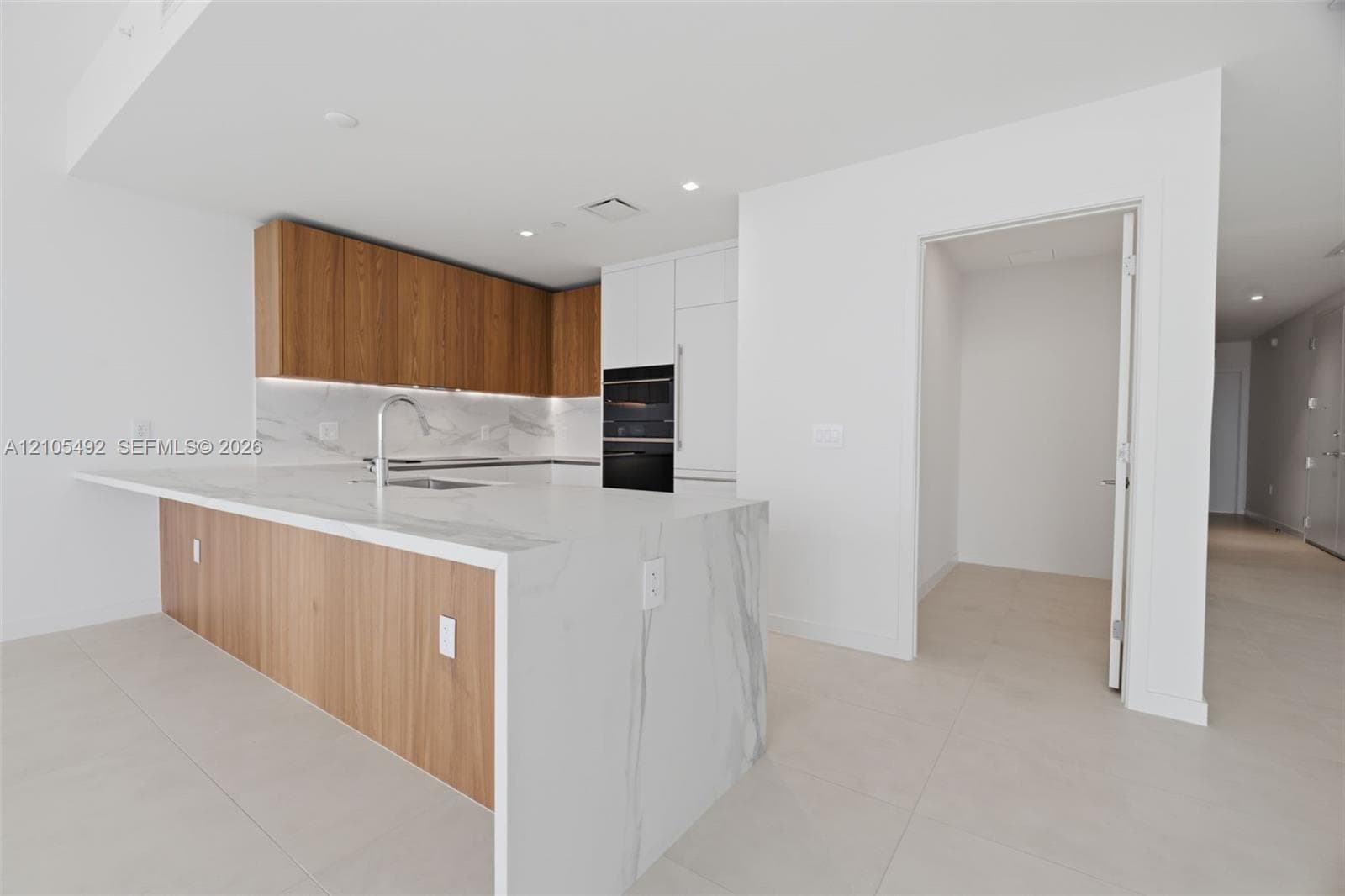 Kitchen featuring wood-finish cabinetry, waterfall-edge stone island, integrated wall oven, neutral-tone large-format floor tile, and under-cabinet lighting