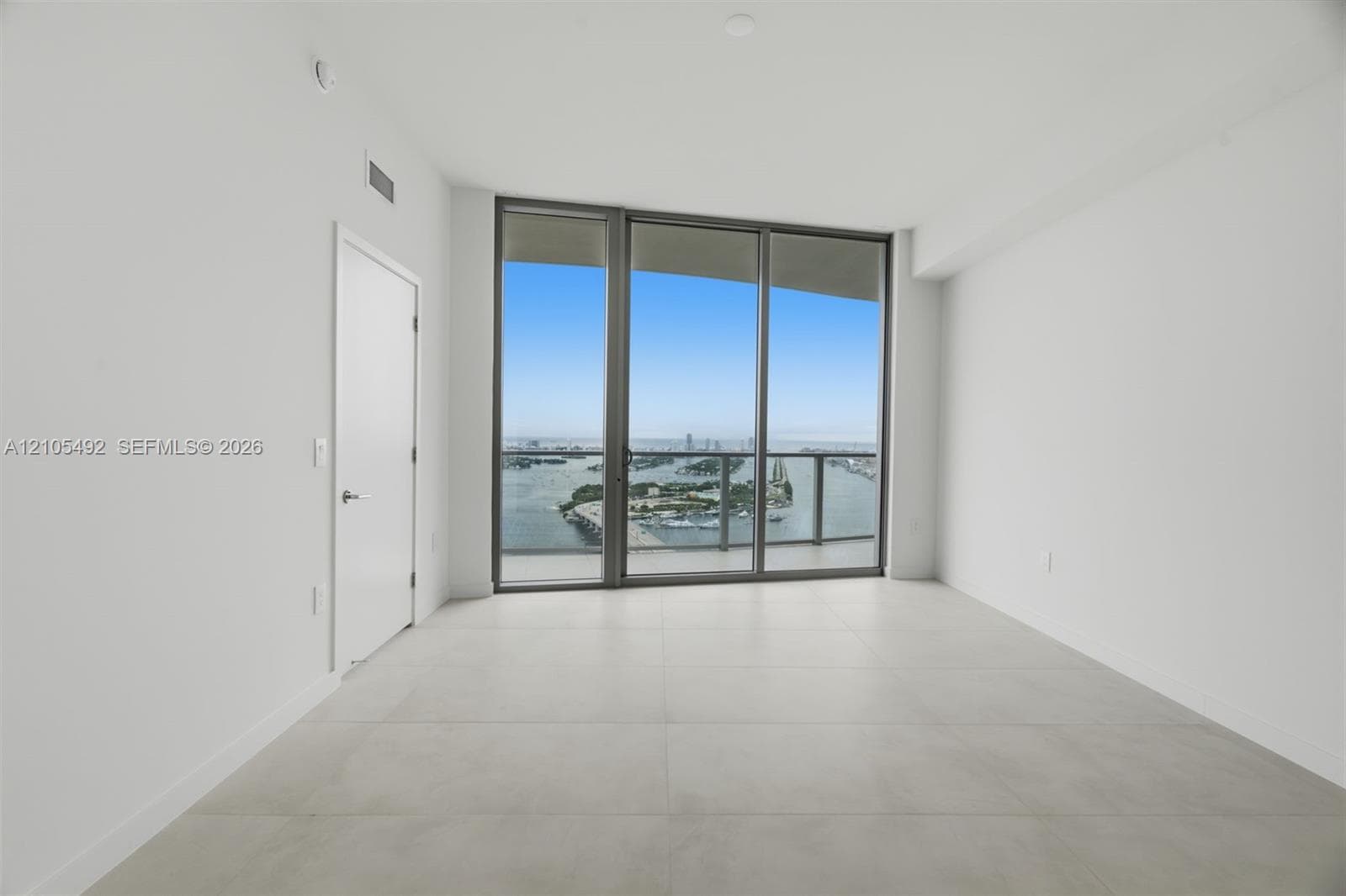 Floor-to-ceiling glass wall, large format tile flooring, and metal-framed balcony access with water and city skyline surroundings