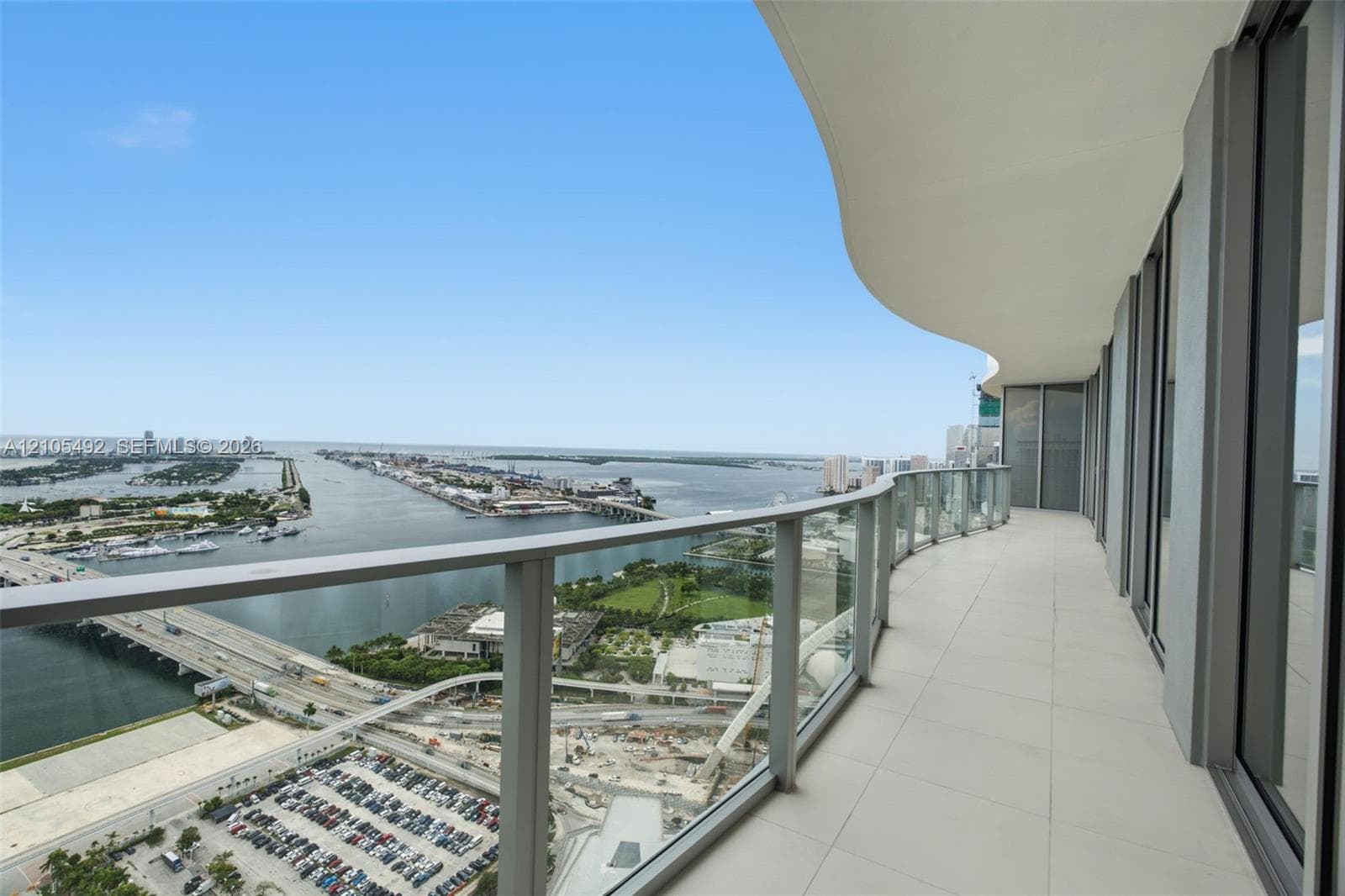 Curved private balcony featuring oversized porcelain tile flooring, glass panel railings, and expansive water and urban scenery