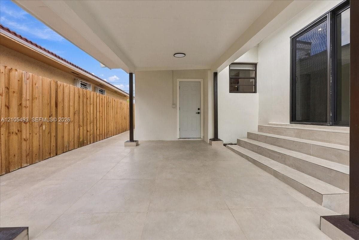 Covered patio featuring large-format porcelain tile flooring, structural support columns, a paneled exterior entry door, and a set of tiered exterior stairs leading to a glass sliding door