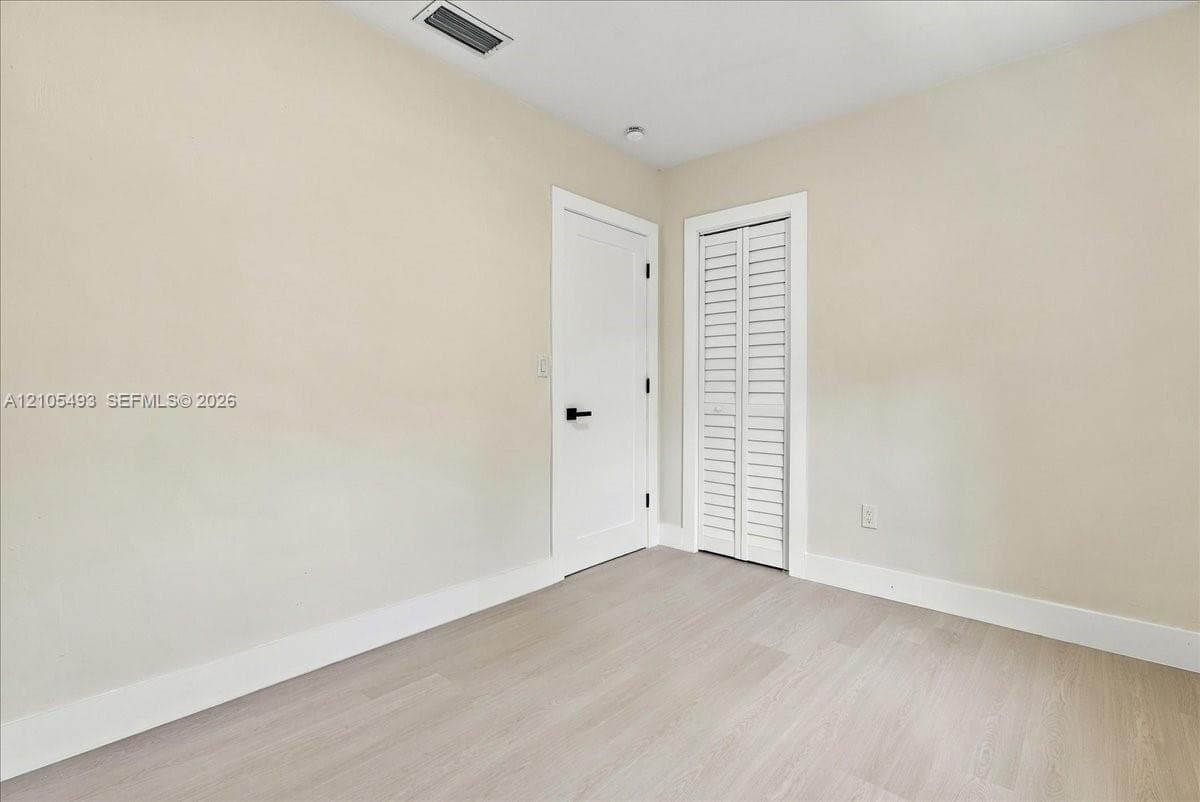 Neutral-toned interior space featuring light wood-finish flooring, a solid-panel door with matte black hardware, and a bifold closet door with louvered panels