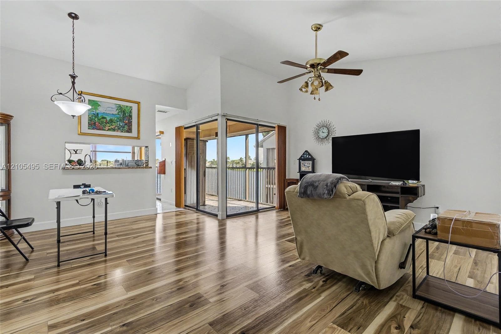 Living area featuring wood-finish flooring, vaulted ceilings, a ceiling fan with integrated light fixture, a pass-through opening, and sliding glass doors with access to an exterior screened balcony