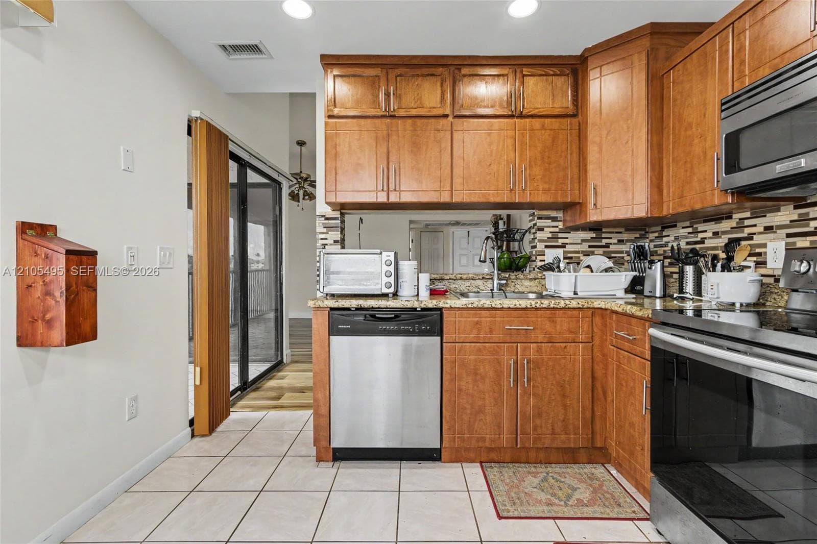 Kitchen featuring stained wood cabinetry, multi-tone tile backsplash, stone-finish countertops, stainless steel dishwasher, and ceramic tile flooring