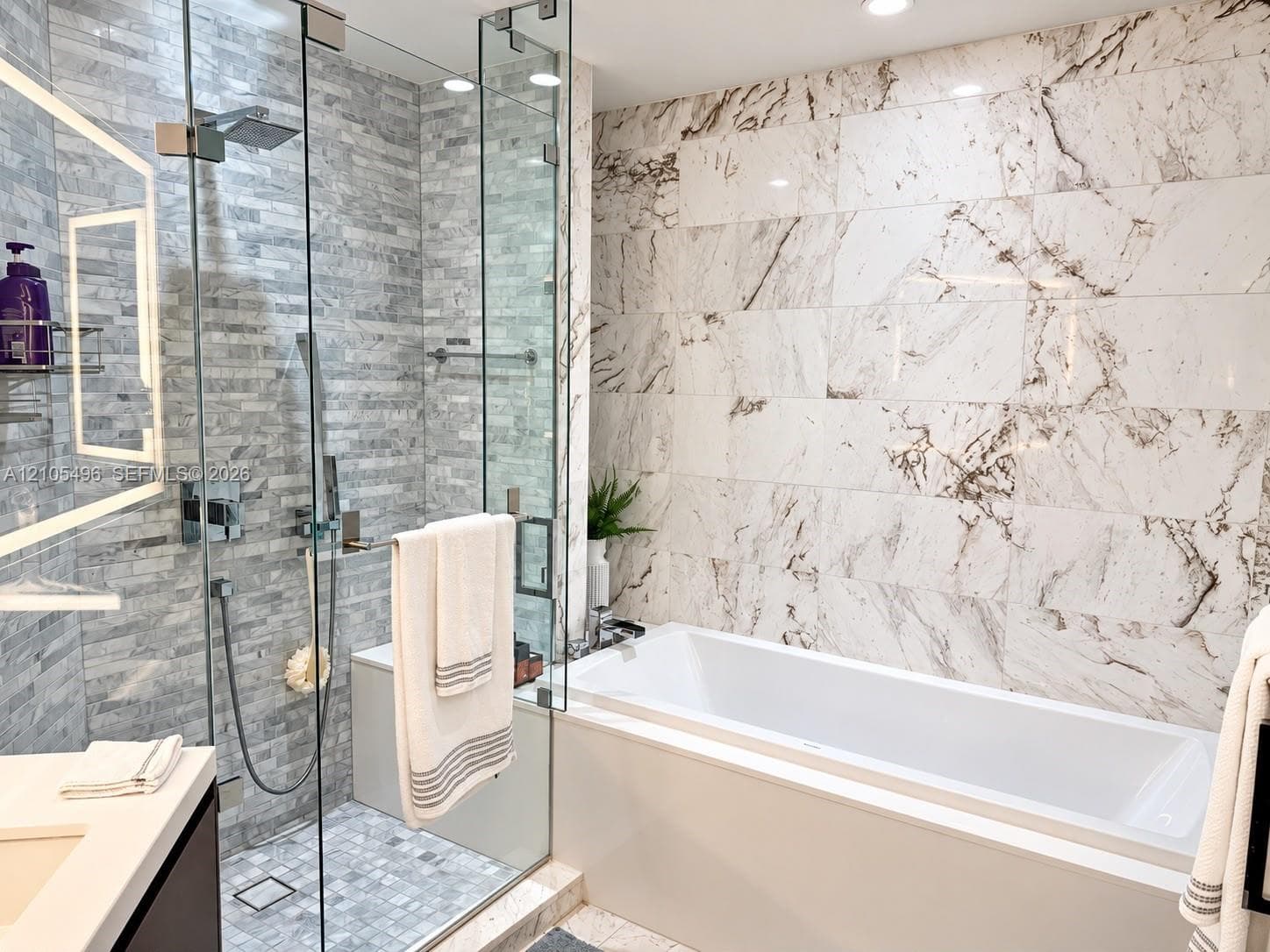 Walk-in glass-enclosed shower with stacked stone tiling, built-in soaking tub featuring a decorative stone-patterned wall surround, and a backlit vanity mirror