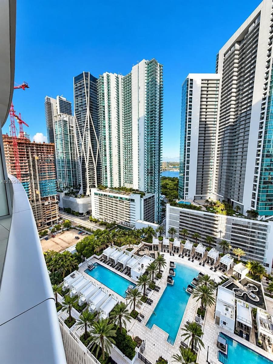 Curved glass balcony railing overlooking an expansive multi-level pool deck with integrated cabanas, palm landscaping, and surrounding metropolitan high-rise architecture