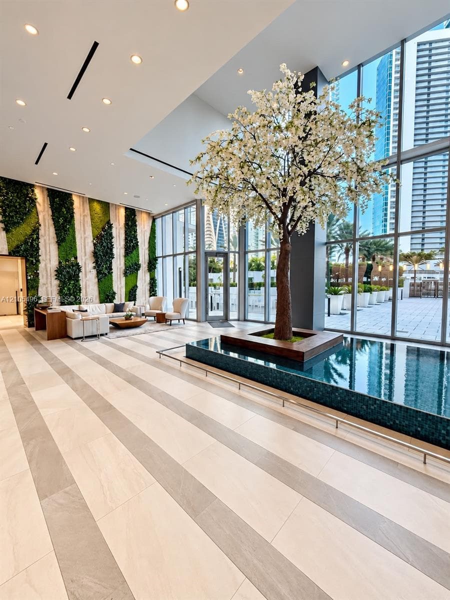 Double-height lobby featuring floor-to-ceiling glass walls, an indoor reflecting pool, a central ornamental tree, and a vertical green wall installation