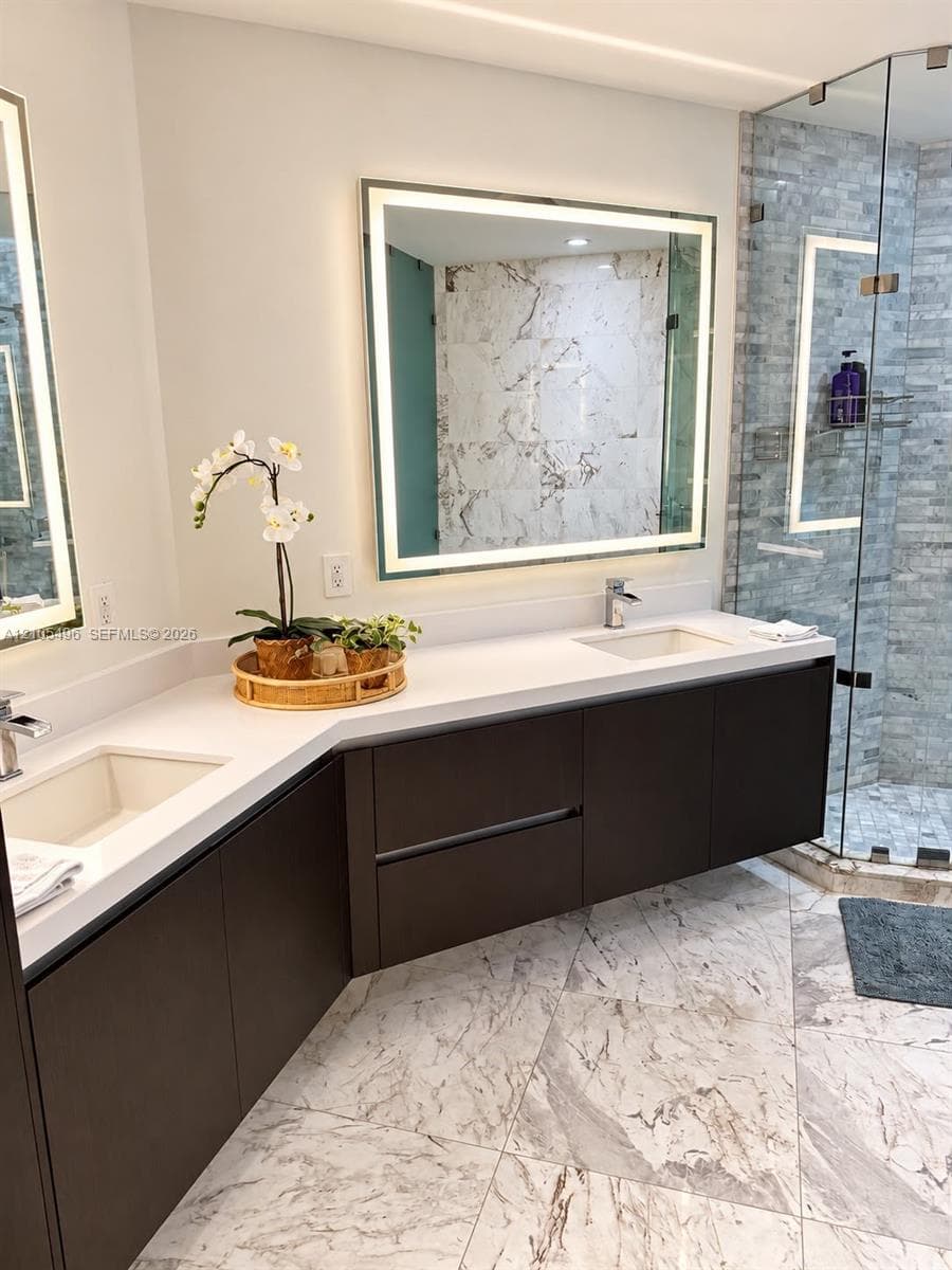 Floating dual vanity featuring dark cabinetry, solid-surface countertops, integrated rectangular sinks, and large, illuminated mirrors, complete with a glass-enclosed shower featuring stone-tiled walls and patterned stone tile flooring