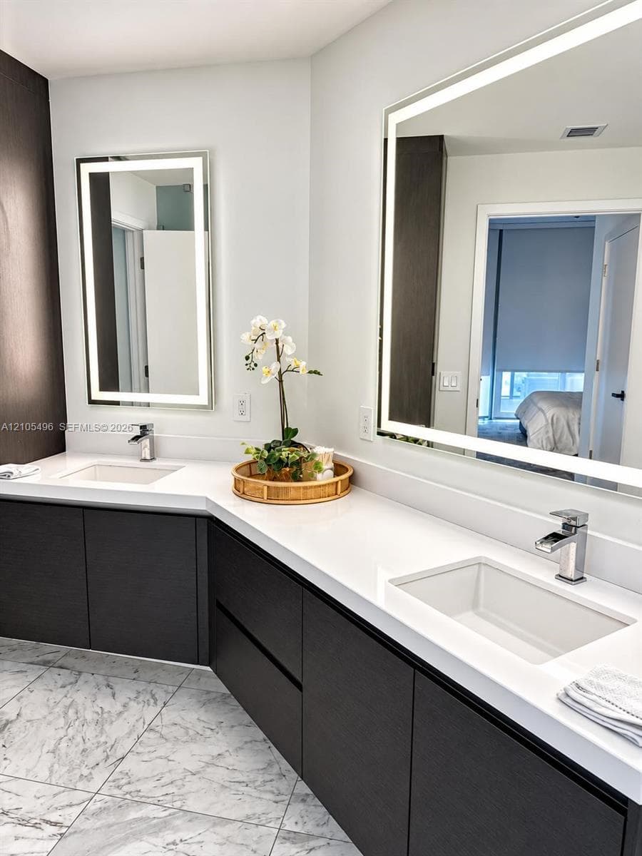 Dual-vanity bathroom featuring dark wood-finish cabinetry, white stone-composite countertops, integrated rectangular sinks with polished chrome faucets, and large rectangular backlit mirrors