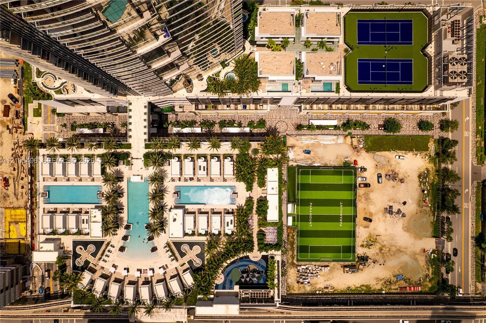 Aerial perspective of residential complex amenities featuring dual swimming pools, two hard-court tennis courts, a turf soccer field, and landscaped courtyards