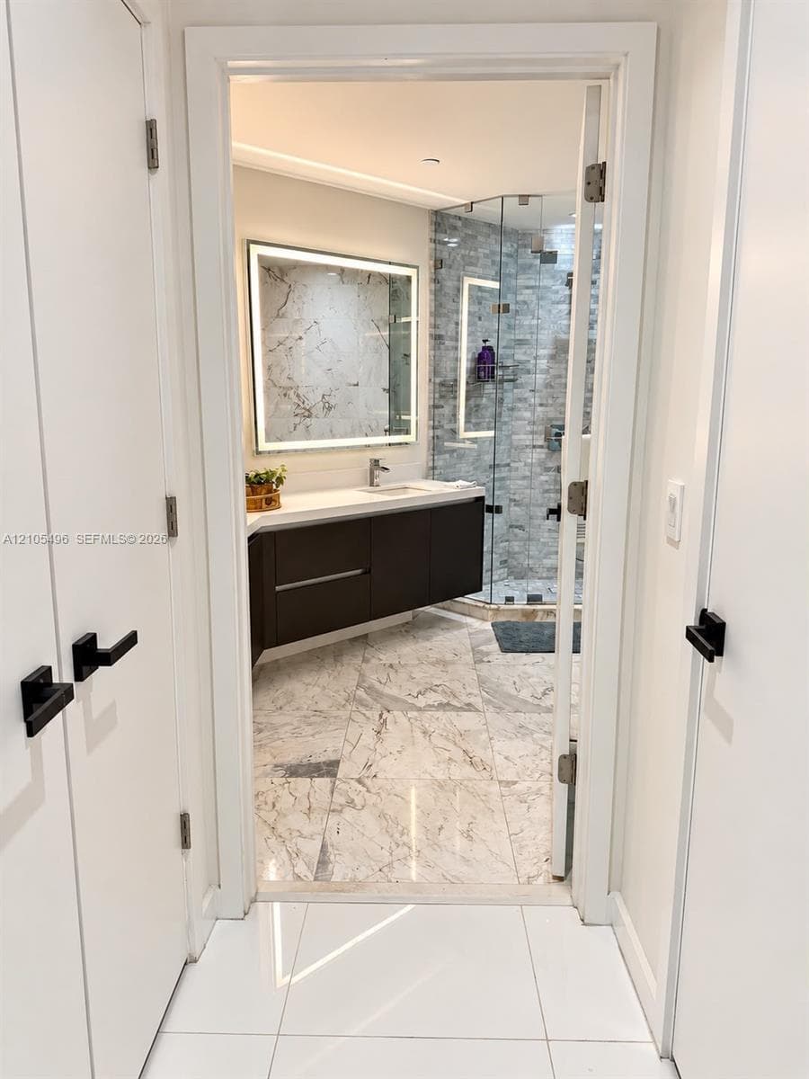 Floating vanity with solid-surface countertop, LED-backlit mirror, glass-enclosed shower with tiled surround, and large-format stone-finish flooring