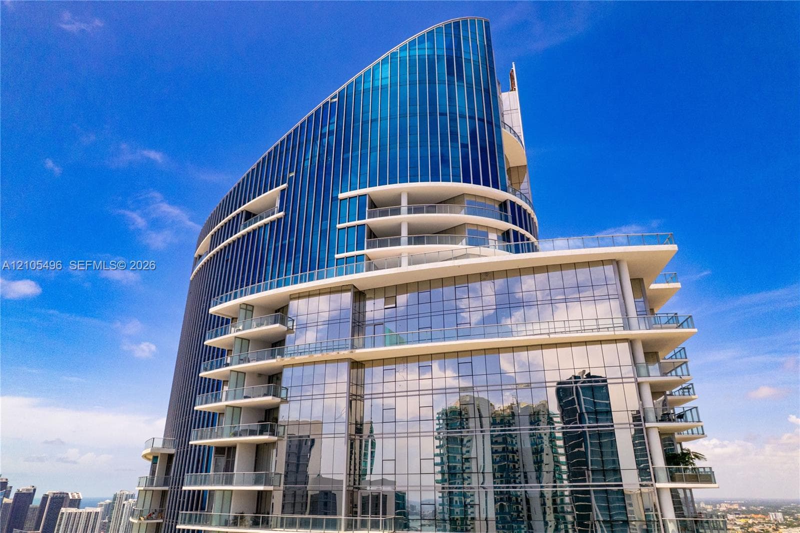 High-rise architectural exterior featuring a curved glass facade, floor-to-ceiling windows, and multiple tiered cantilevered balconies with glass railings