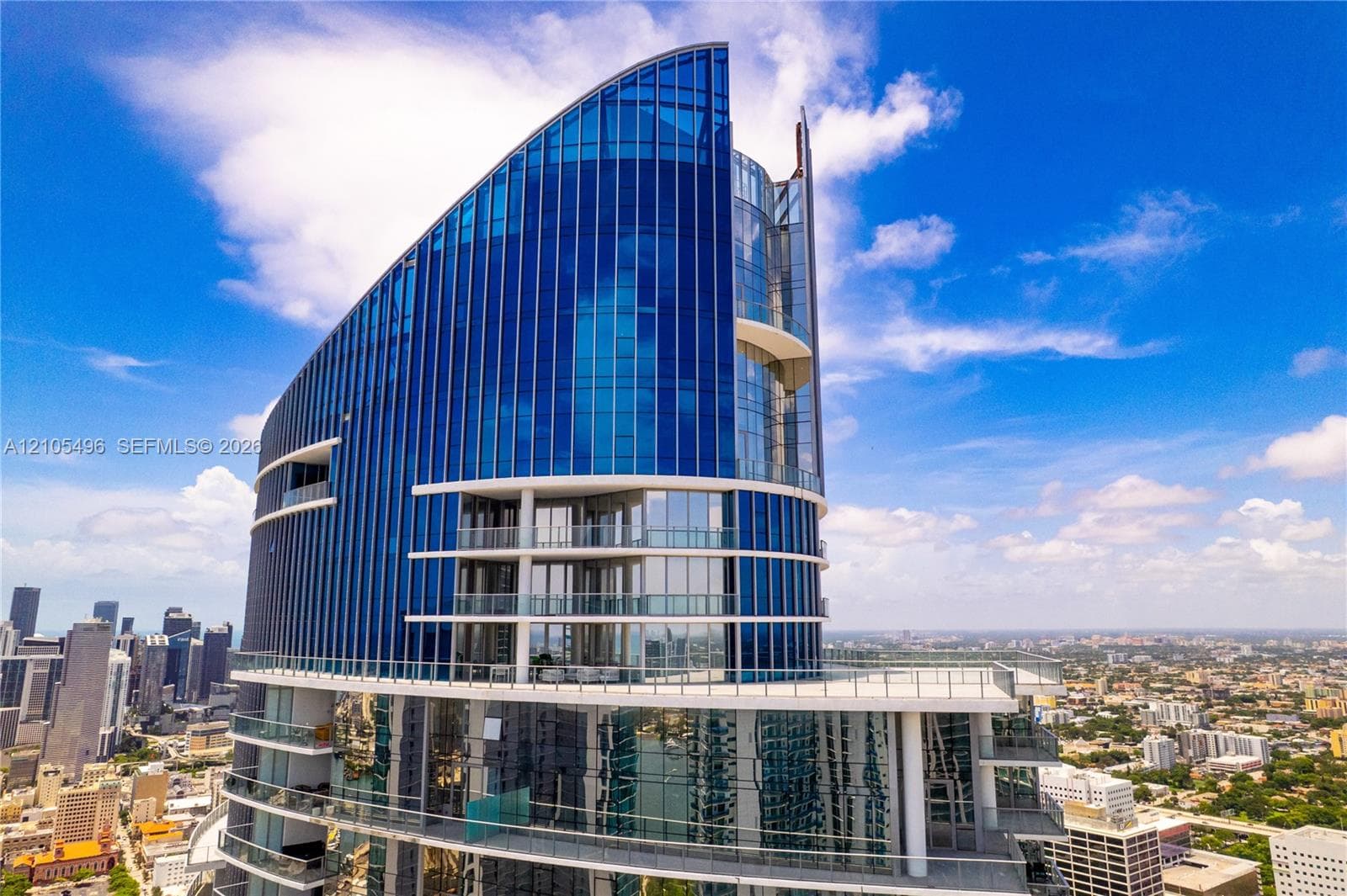 High-rise architectural design featuring a curved blue glass curtain wall, multiple levels of wraparound balconies, and metal railings