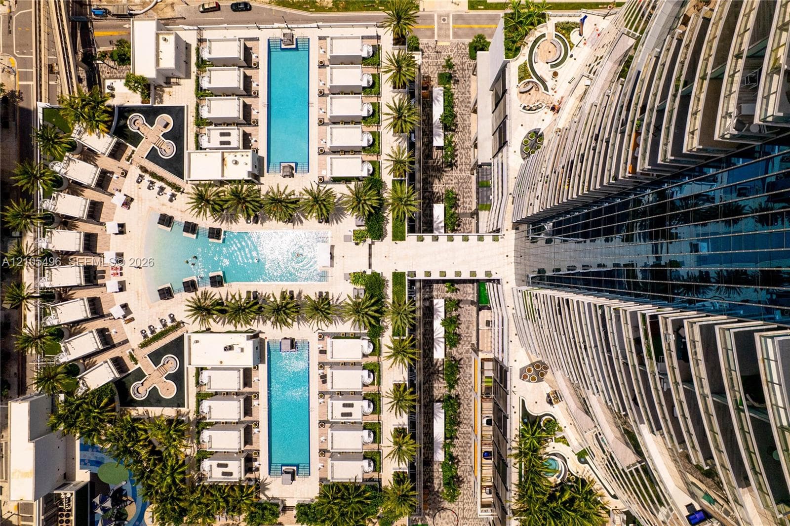 Aerial view of a high-rise residential structure featuring multi-level swimming pools, paved sun decks, palm tree landscaping, and cabana structures
