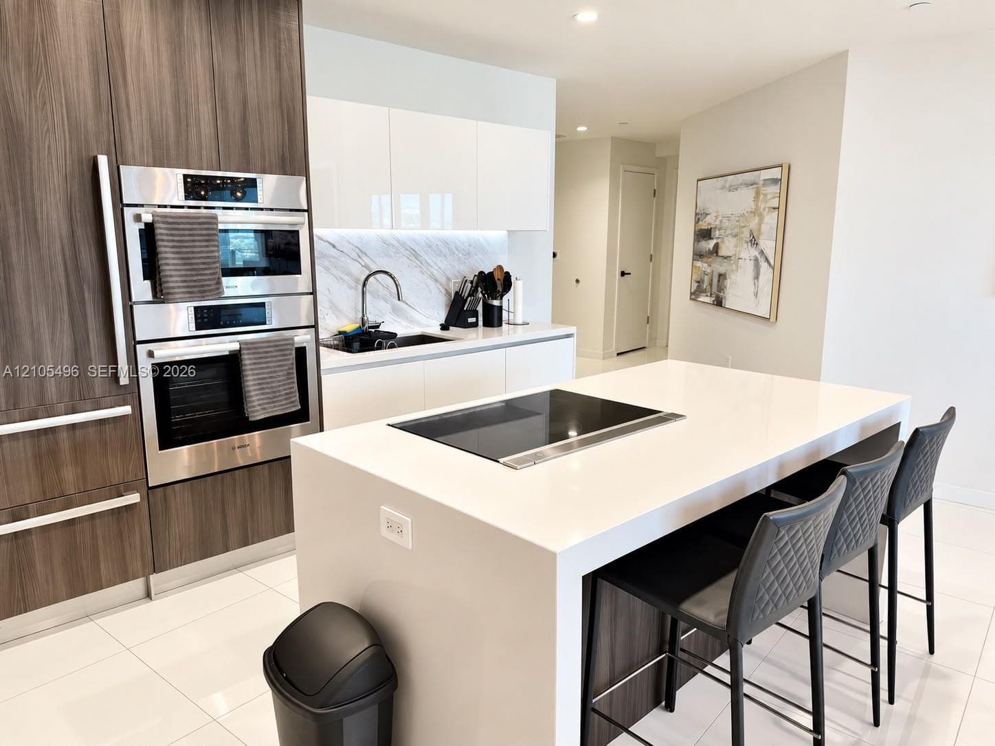 Kitchen featuring high-gloss white cabinetry, wood-finish floor-to-ceiling pantry units, a large quartz-topped island with an integrated induction cooktop, and stone-finish backsplash