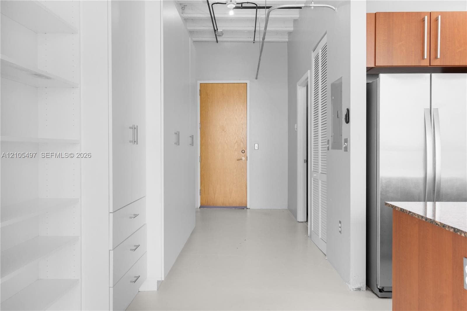 White built-in cabinetry with silver-tone hardware, wood-panel entry door, light-toned flooring, exposed ceiling conduit, and a stainless steel refrigerator