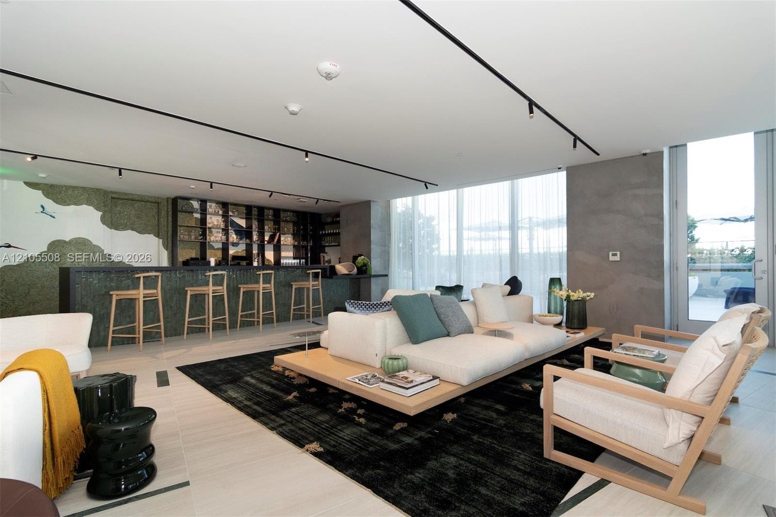 Integrated wet bar with barstool seating, recessed track lighting, full-height windows with sheer window treatments, concrete accent wall, and light-toned flooring