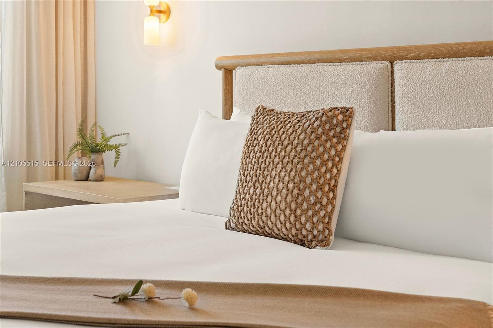 Custom wood-framed headboard with textured upholstery, light-toned wood-finish side table, and wall-mounted brass light fixture with frosted glass