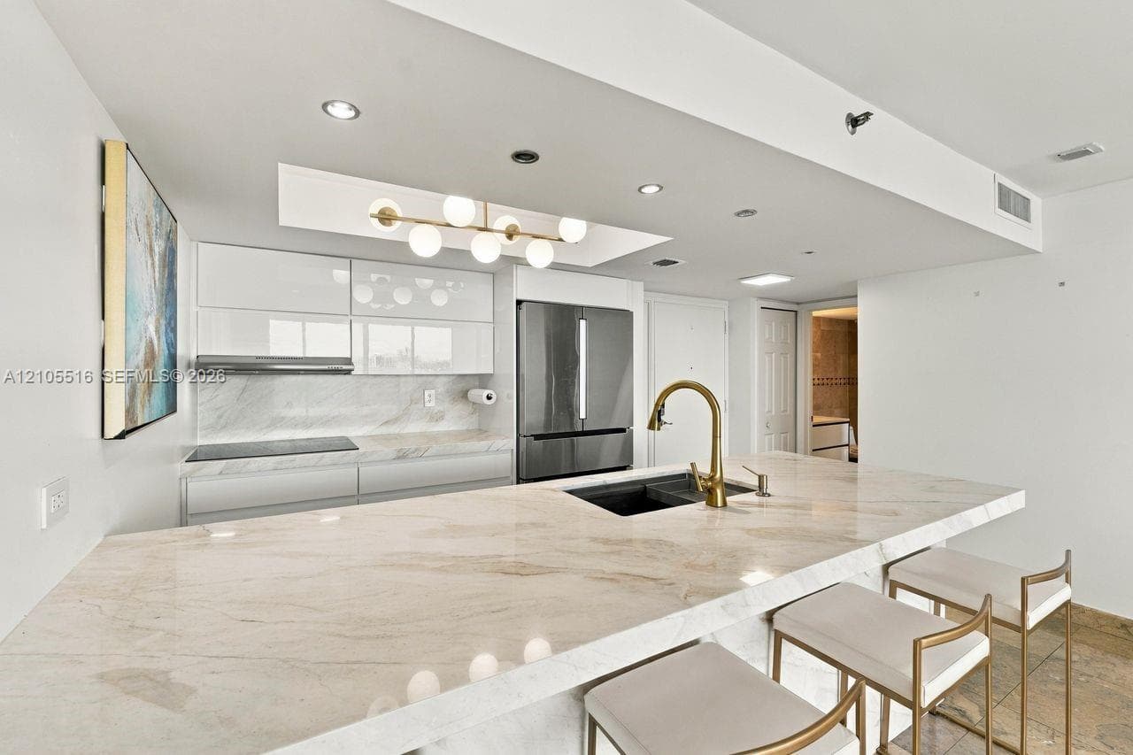 Extended kitchen island with stone countertops, modern pendant lighting, integrated sink with a gold-tone faucet, and white glossy cabinetry
