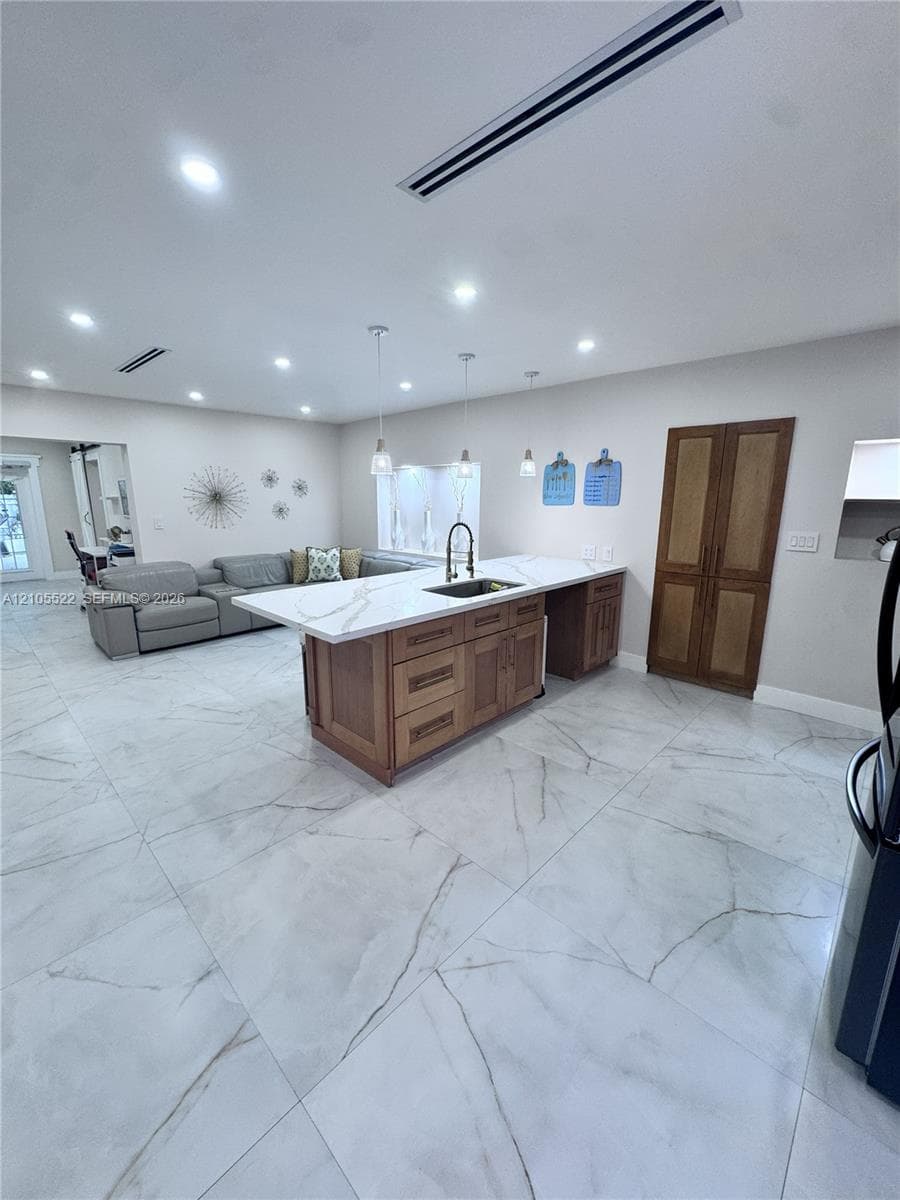 Spacious open-concept room featuring large format tile flooring, a kitchen island with a white countertop and integrated sink, pendant lighting, recessed ceiling lights, and wooden double doors