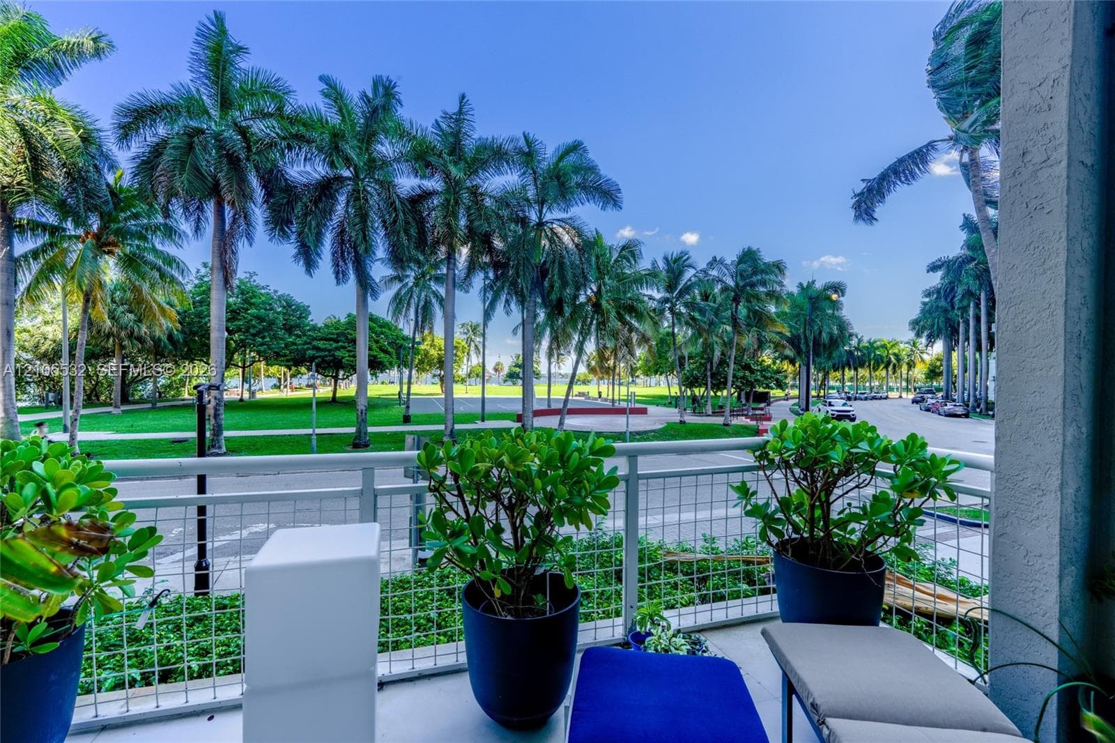 Covered balcony featuring white metal railings and concrete flooring, overlooking a green space with mature palm trees