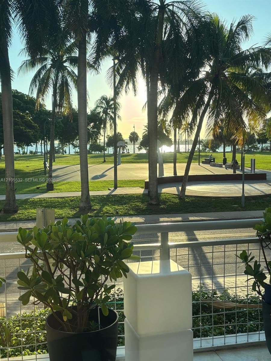 Balcony with a wire-mesh railing overlooking a public park featuring a paved sports court, paved pathways, and mature palm trees