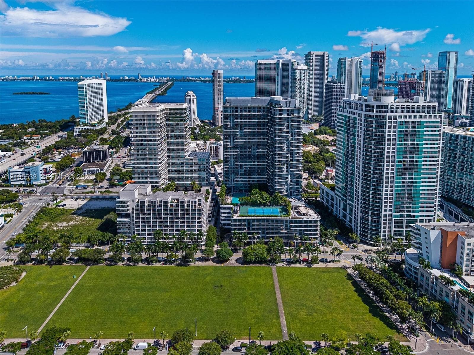 Aerial perspective of high-rise urban development featuring a rooftop swimming pool and recreational court, situated in proximity to coastal waterways